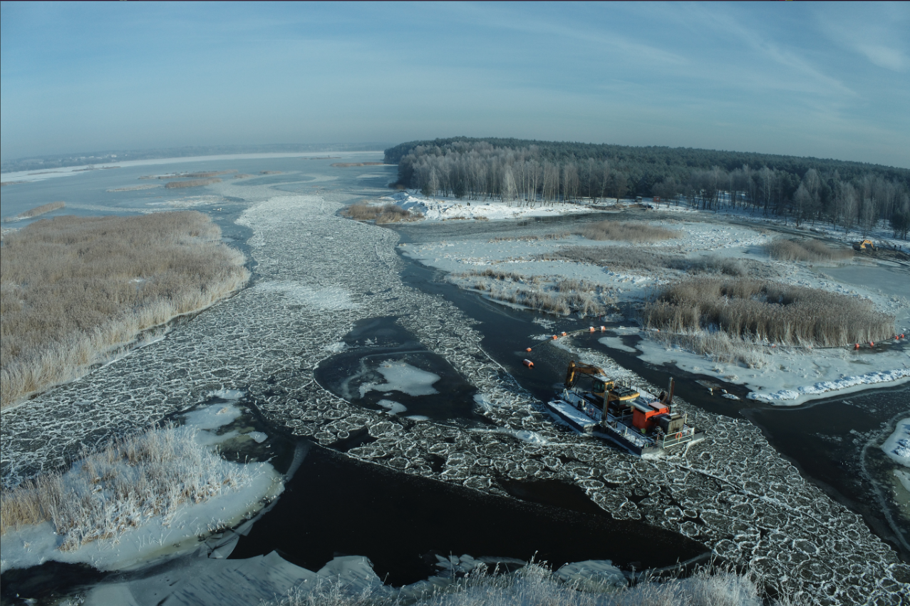 Widok z lotu ptaka na specjalistyczną platformę hydrotechniczną z ramieniem koparki, pracującą przy udrażnianiu szlaku wodnego i kruszeniu pancerzy lodowych w zimowym krajobrazie rzeki, w tle ośnieżone trzciny i las.